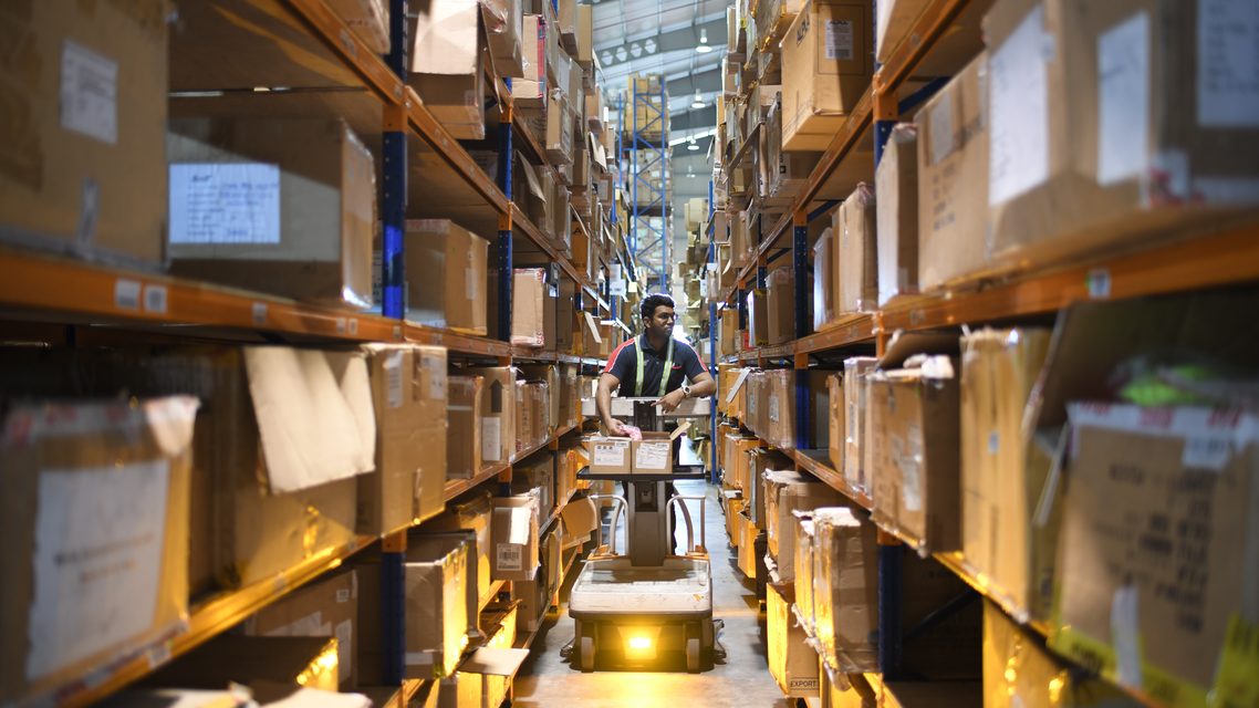 An operator bringing a powered pallet truck down a racking aisle, cartons stacked to the roof on both sides.