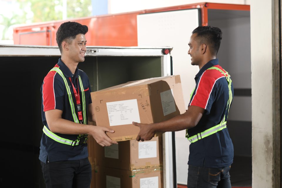 Two colleagues handing a carton between them at the tail of a lorry, pallets loaded behind.