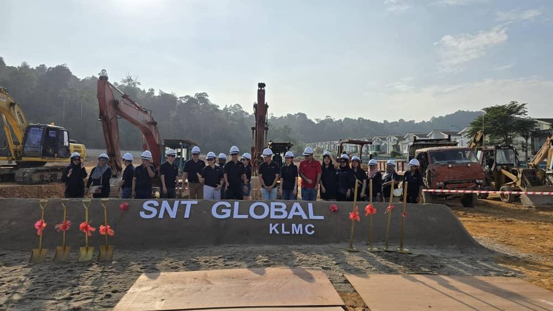The SNT Global KLMC groundbreaking: the team in hard hats behind a bank of earth carrying the site name, excavators and ceremonial spades on either side.