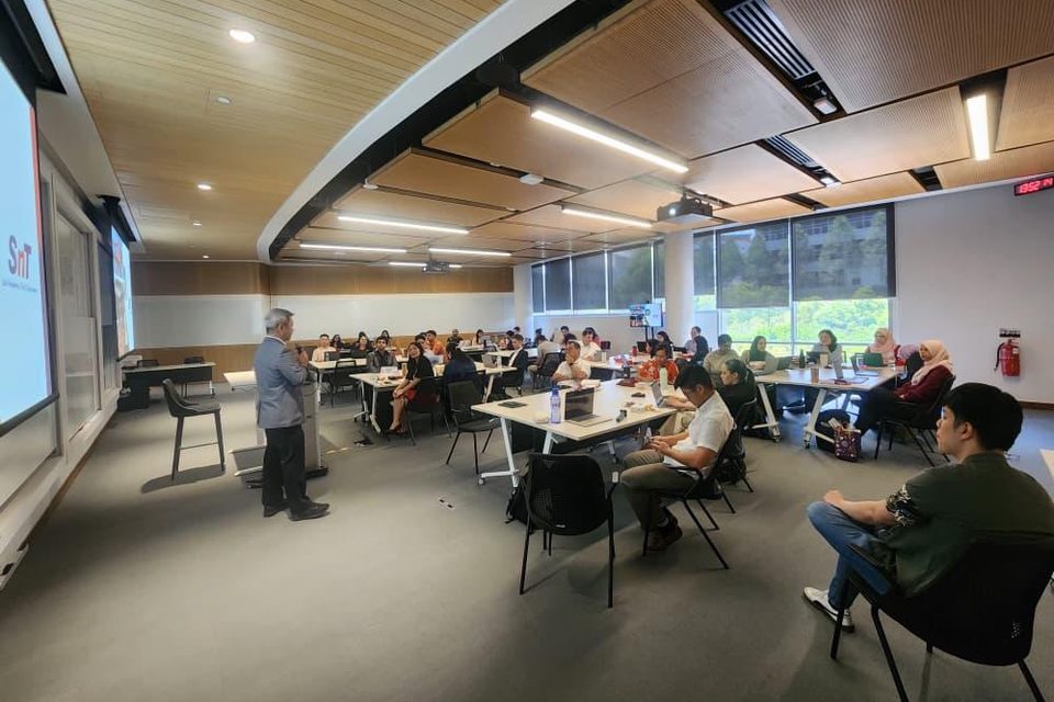 A session in progress in a tiered classroom, an SNT slide on the screen and the room seated at tables.