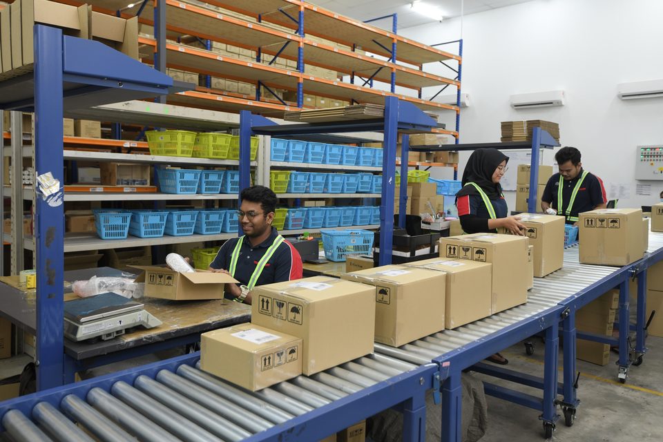 Three staff working a pack bench, cartons moving down a roller conveyor and picking totes racked behind them.