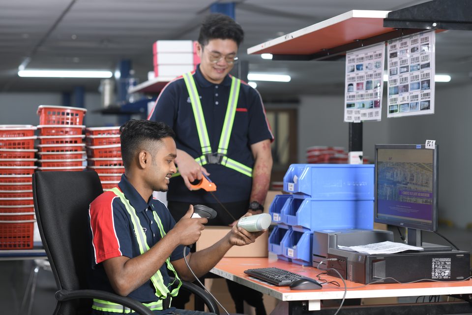 A supervisor standing over a seated colleague at a scanning station, both looking at the item being scanned.