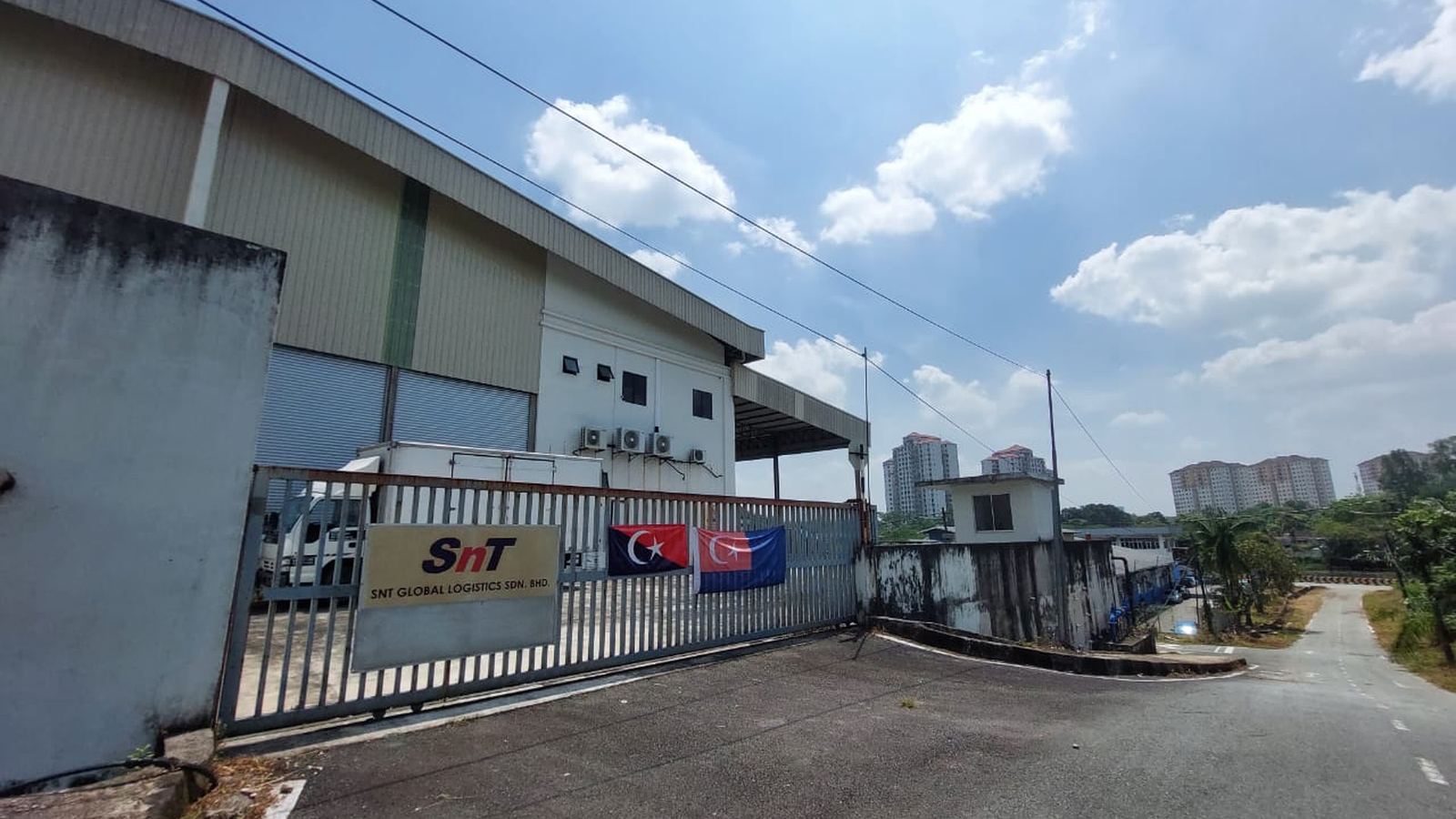 The gated entrance to the Johor Bahru facility, the SNT Global Logistics sign on the fence with the Malaysian and Singaporean flags beside it.
