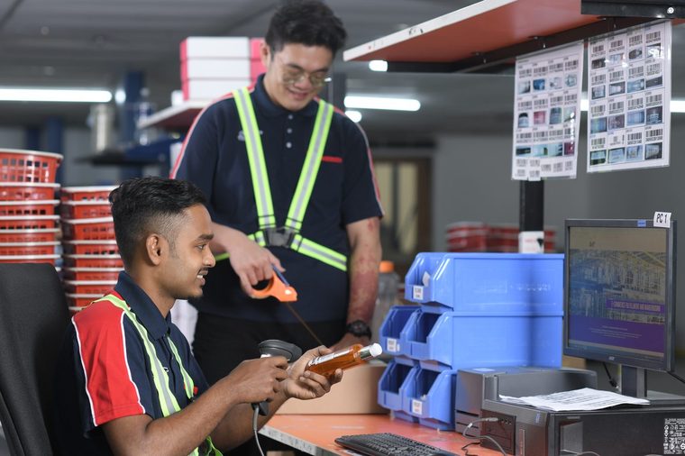 Two colleagues at a goods-in workstation, one scanning an item beside a monitor and stock bins.