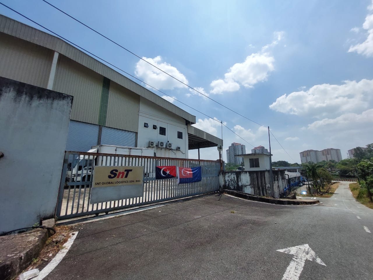 The gated entrance to the Johor Bahru facility, the SNT Global Logistics sign on the fence and Johor state flags beside it.